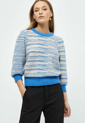 Niebieski sweter w paski z okrągłym dekoltem, miękką tkaniną i rękawami o długości 3/4; dopasowane niebieskie ściągacze na dole i na rękawach. Połączony z czarnymi spodniami.