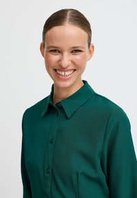 Smaragdgrünes Langarmhemd mit Knopfleiste, Kragen und glatter Textur. Verfügt über ein figurbetontes Design und einen minimalistischen Stil.