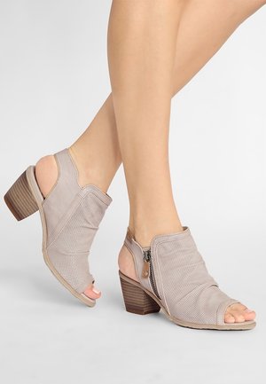 Sandales à talon bloc en cuir beige perforé avec bout ouvert, fermeture éclair latérale et bride arrière, portées sur des jambes croisées sur un fond uni.
