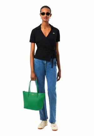 Femme portant un haut portefeuille noir, un jean bleu, des baskets blanches, des lunettes de soleil, tenant un sac fourre-tout vert sur un fond blanc.