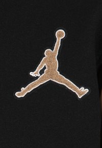 Czarna tkanina z brązowym, teksturowanym haftowanym logo Jumpman, obramowanym na biało, przedstawiającym postać w skoku do koszykówki.