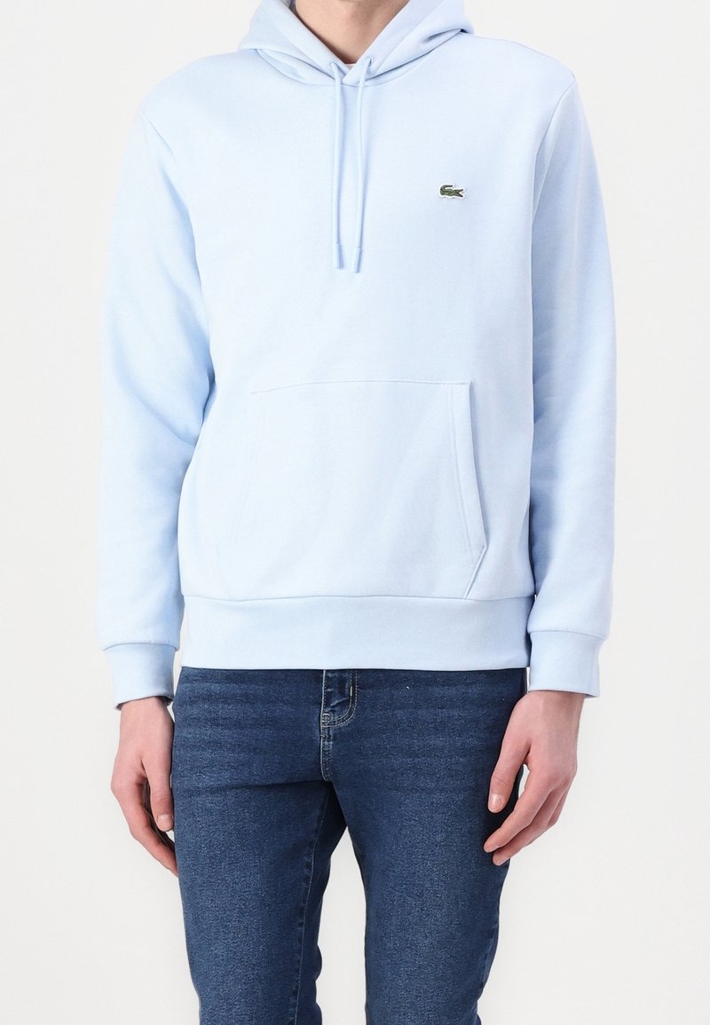 Personne portant un sweat à capuche bleu clair avec une poche avant et un petit logo crocodile vert sur la poitrine, associé à un jean bleu foncé.