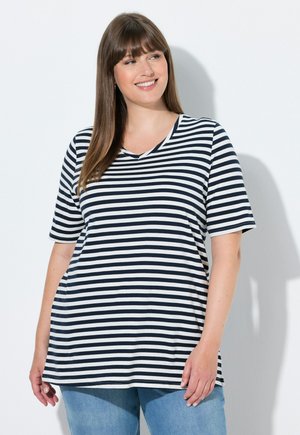 Uśmiechnięta kobieta o prostych brązowych włosach, ubrana w czarno-białą pasiastą koszulkę z krótkim rękawem i jasnoniebieskie dżinsy, stojąca na białym tle.