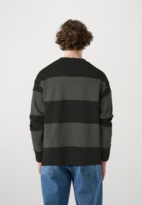 Pikend käised pullover, millel on vahelduvad mustad ja hallid horisontaalsed triibud, ümmargune kaelus ja mugav lõige. Denim teksad on nähtavad talje juures.