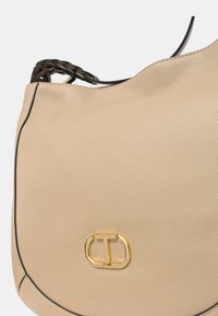 Borsa a mano in pelle beige con superficie texturizzata, bordi neri e chiusura in metallo dorato con un emblema. Dettaglio della maniglia intrecciata.