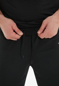 Sorte atletiske bukser med snor i taljen. Glat tekstur med en tætsiddende pasform. Hænder der griber fat om taljen, hvilket fremhæver en minimalisk stil.