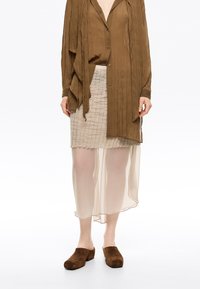 Blusa marrón arrugada con cierre de botones, superpuesta a una falda beige transparente con un patrón texturizado, combinada con zuecos de ante marrones.