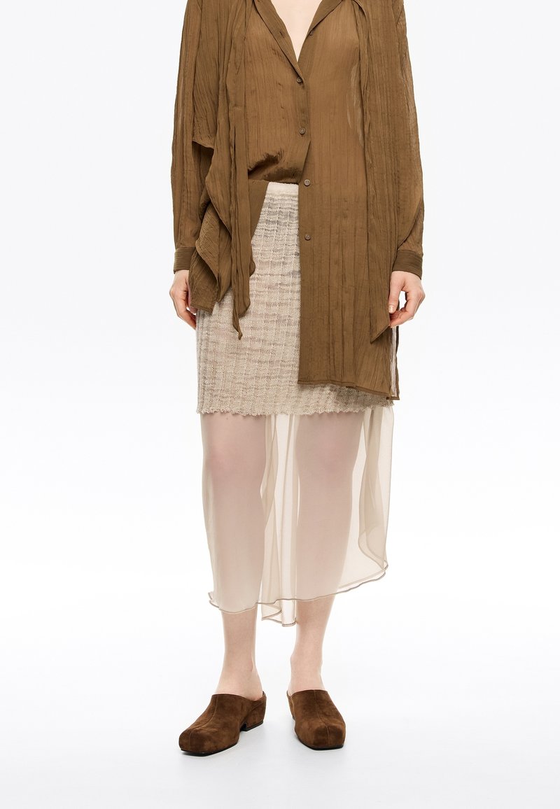 Blusa marrón arrugada con cierre de botones, superpuesta a una falda beige transparente con un patrón texturizado, combinada con zuecos de ante marrones.