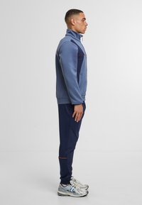 Světle modrá zipová bunda s námořnickými akcenty, v kombinaci s námořnickými kalhotami a stříbrnými sportovními botami. Materiály vypadají hladce a jsou dobře padnoucí.