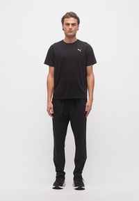 Άνδρας που στέκεται βλέποντας μπροστά, φορώντας μαύρο μπλουζάκι Puma, μαύρο παντελόνι και μαύρα αθλητικά παπούτσια, μπροστά σε απλό λευκό φόντο.