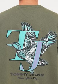 Olivengrøn T-shirt med et stort grafisk tryk af en flyvende and og bogstaverne "TJ" i teal, med "TOMMY JEANS New York City" trykt nedenfor.