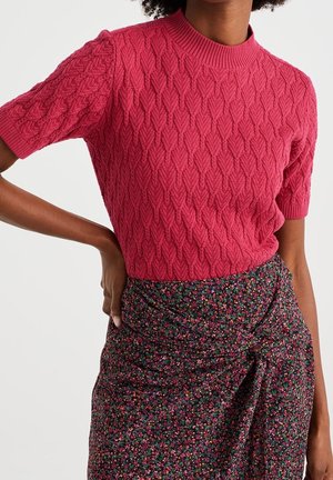 Kobieta w różowym, krótko rękawowym, dzianinowym topie o teksturowanej powierzchni oraz wysokiej taliowanej, ciemnej, kwiatowej spódnicy z detalem w postaci węzła, z ręką opartą na biodrze.