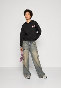 Žena u crnoj majici s kapuljačom s ružičastim leptirima, širokim blijedim cargo trapericama, srebrnim tenisicama i s malom ružičastom torbicom stoji nasuprot bijeloj pozadini.