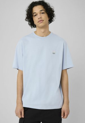 Jeune homme aux cheveux bouclés portant un t-shirt bleu clair oversize et un pantalon noir sur un fond gris uni.