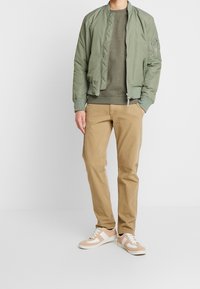 Chaqueta bomber acolchada verde, sudadera oliva, pantalones chinos beige y zapatillas beige y blancas. Diseño entallado con puños y cinturilla de canalé.