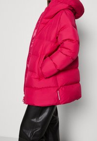 Chaqueta acolchada rosa con capucha, con cierre de cremallera, bolsillos laterales y puños elásticos. Combinada con pantalones de cuero negros.