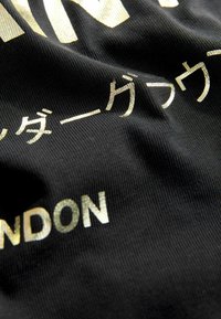 Melna auduma ar spīdīgu zelta tekstu. Dizainā ir japāņu rakstzīmes un "LONDON" izceltā burtveidā. Redzam teksturētu virsmu.