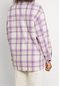 En person som bär en skjorta med lavendel- och beige-rutigt mönster med långa ärmar och vitaktiga byxor, sedd bakifrån mot en enfärgad bakgrund.