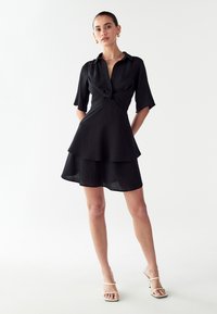 Vestido negro con falda en capas, mangas cortas, un frontal anudado y un cuello. Textura suave y tejido ligero.