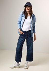 Světle modrá zipová bunda, bílý tričko, tmavé džíny s širokými nohavicemi a bílé tenisky s světle modrými akcenty a kšiltovka s logem.