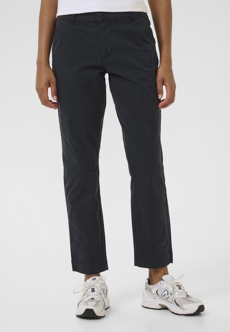 Personne portant un pantalon droit noir et des baskets blanches New Balance, debout sur un fond clair uni.