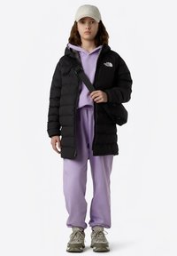 Casaco acolchoado preto com capuz, usado sobre uma sweat-shirt roxa clara e calças de fato de treino combinando. Ténis cinzentos com solas texturizadas. Boné bege.