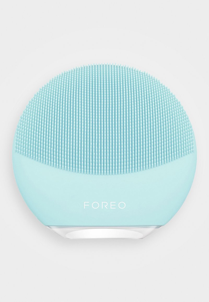 Ansiktsrensingsenhet, rund form, lyseblå silikonmateriale, teksturert med små hevede prikker, med en hvit bunn og merking.