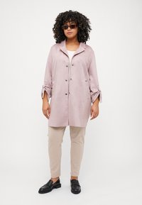 Light pink suede coat with a collar, two pockets, and button closure, paired with beige pants and black loafers. Textured fabric, relaxed fit.