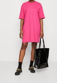 Vaaleanpunainen puuvillainen t-paitamekko, jossa on lyhyet hihat, rento leikkaus ja pieni logoyksityiskohta. Yhdistetty mustien lakatut loafereitten ja kiiltävän olkalaukun kanssa.