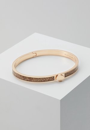 Złocona bransoletka typu bangle z błyszczącym brązowym inkrustowanym wzorem i okrągłym zapięciem, umieszczona na białym geometrycznym piedestale.