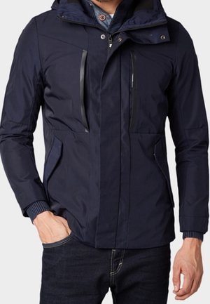 Mężczyzna w granatowej kurtce z kapturem, z zamykanymi na zamek kieszeniami na piersi i po bokach, ubrany w ciemne dżinsy.