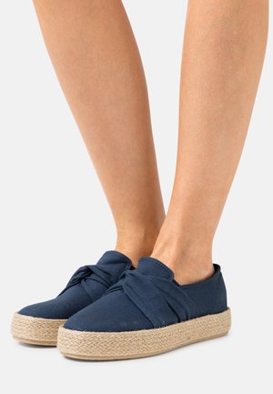 Espadrilles en toile marine avec un grand design noué, dotées d'une semelle en corde de jute. Texture lisse avec un design plat et une pointe ronde.