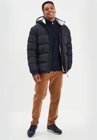 Schwarze gepolsterte Jacke mit fleecegefütterter Kapuze, marineblauer Pullover, braune Cordhosen und dunkle Sneakers, mit durchgehendem Reißverschluss und sichtbaren Nähten.