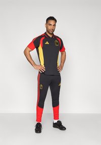 Conjunto desportivo preto e vermelho composto por uma camisola tipo polo com detalhes amarelos e logotipo, acompanhado por calças ajustadas com seções vermelhas e riscas.