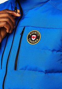 Blaue gepolsterte Jacke mit einer Reißverschluss-Tasche und einem runden Aufnäher mit dem Text "ZAVETTI" und "KANADA" sowie einem roten Ahornblatt-Detail.