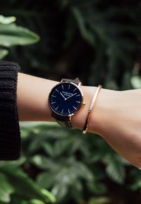Reloj redondo con caja de oro, cara negra y marcadores de hora blancos, acompañado de una fina pulsera de oro en una muñeca, con un fondo de follaje verde.
