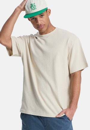 Beige strukturiertes T-Shirt mit kurzen Ärmeln, Rundhalsausschnitt und lockerem Schnitt. Kombiniert mit einer weißen Kappe mit grünen Akzenten und Design.