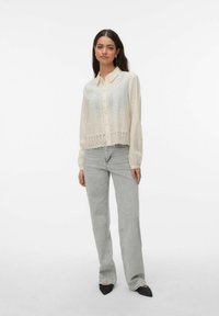 Femme debout portant une blouse crème à boutons avec des détails en dentelle et un jean gris clair à jambes droites avec des chaussures noires à bout pointu.