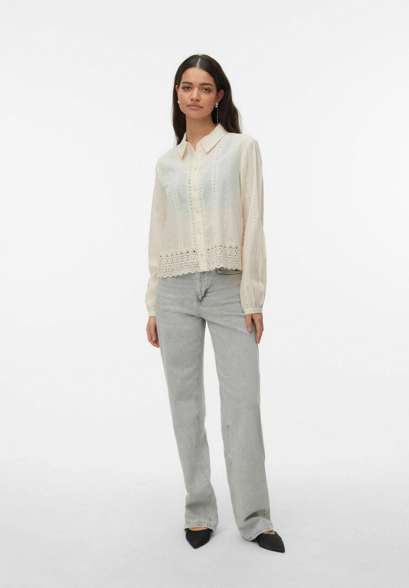 Femme debout portant une blouse crème à boutons avec des détails en dentelle et un jean gris clair à jambes droites avec des chaussures noires à bout pointu.