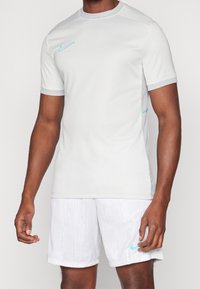 Šedé sportovní tričko s krátkými rukávy, kulatým límcem a tyrkysovým logem Nike. V kombinaci s bílými šortkami se vzorem.