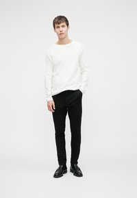 Bela dolgih rokavov srajca, črne ozke hlače in črne čevlje. Preprost dizajn, teksturirana tkanina in nevtralna barvna paleta.