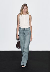 Top senza maniche beige chiaro, jeans a gamba larga blu sbiaditi, sandali neri con tacco, tiene una borsa marrone scuro con una chiusura in argento.