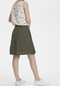 Hvid ærmeløs top med blomstermønster, parret med en olivengrøn nederdel i knæ-længde. Modellen har hvide sneakers på. Blødt stof, løs pasform.