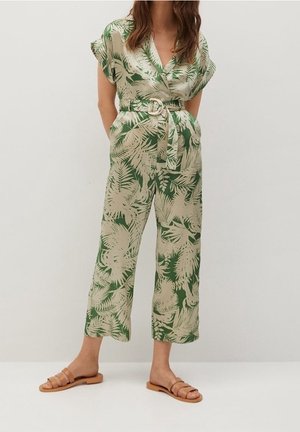 Femme portant une combinaison ceinturée imprimée de feuilles tropicales vertes et beiges, avec des manches courtes et des sandales beige, se tenant devant un fond uni.