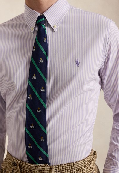 Muž nosi svetlofialovú pruhovanú košeľu s gombíkmi, tmavomodro-zelenú pruhovanú kravatu a hnedé kockované nohavice.