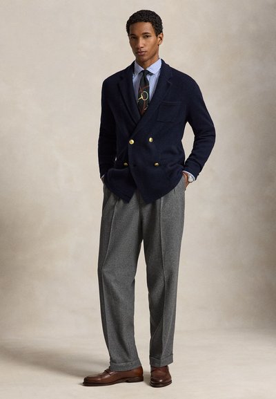 Uomo in piedi con le mani nelle tasche che indossa un blazer blu navy con bottoni dorati, camicia a righe, cravatta fantasia, pantaloni grigi e scarpe di pelle marrone.