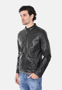 Giacca in pelle nera con colletto alla coreana, zip frontale e due tasche laterali. Tessuto liscio e vestibilità su misura. Indossata sopra una maglietta grigia.