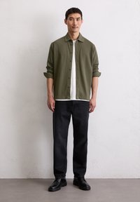 Camicia verde oliva con bottoni sopra una t-shirt bianca, abbinata a jeans neri a gamba dritta e scarpe nere stringate. Design minimalista, vestibilità casual.