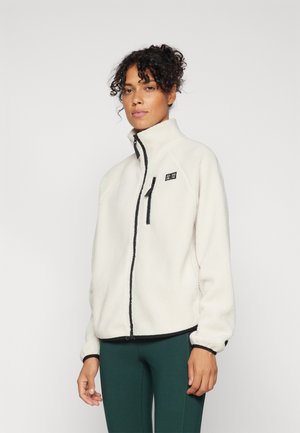 Femme aux cheveux bouclés portant une veste polaire blanche avec des détails de fermeture éclair noirs et un pantalon vert foncé sur un fond clair uni.