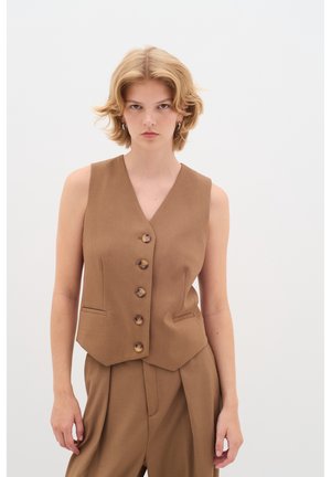 Junge Person mit kurzen blonden Haaren, die eine braune, knöpfbare Weste und passende weite Hosen trägt, vor einem schlichten weißen Hintergrund.